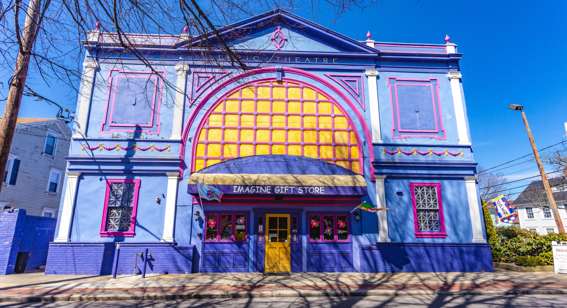 Front of Imagine Gift Store. A building with different shades of purple that is labeled "Lyric theater" at the top. There is a large mock window painted yellow with magenta highlights. Flags wave out front.