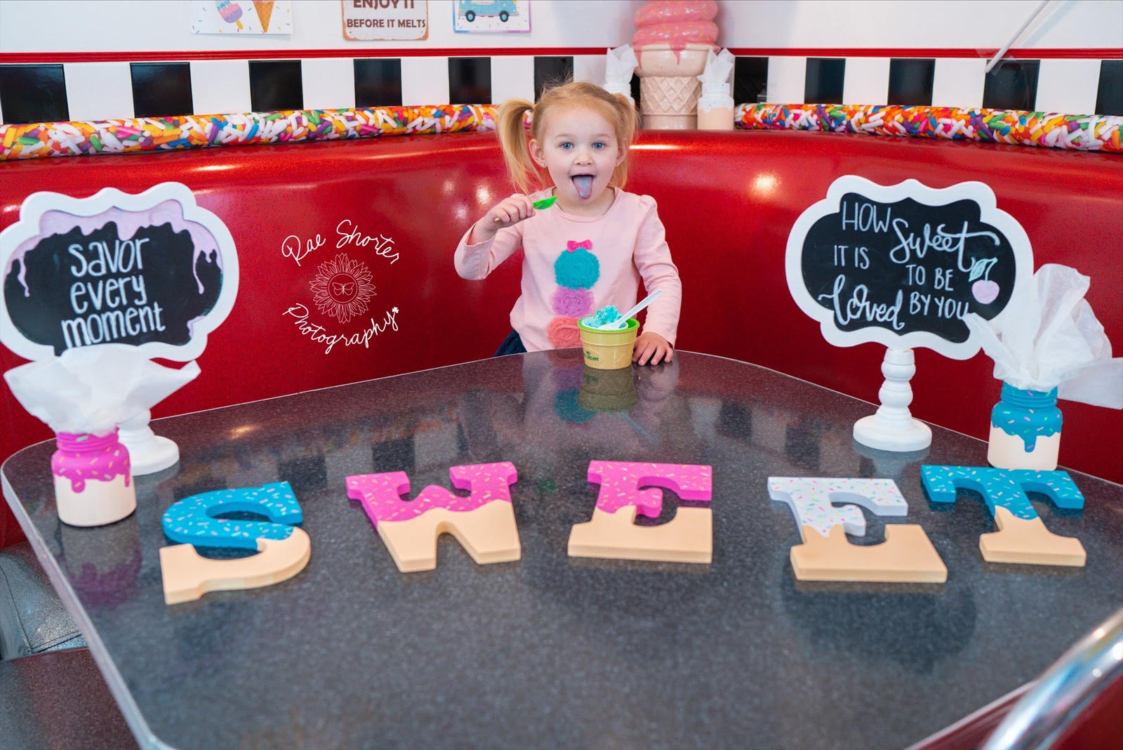 A girl poses in a both of an ice cream parlor with the letters that spell "Sweet" laid out on the table in front of her. Signs that say "Savor evey moment" and "How sweet it is to be loved by you" are next to her.
