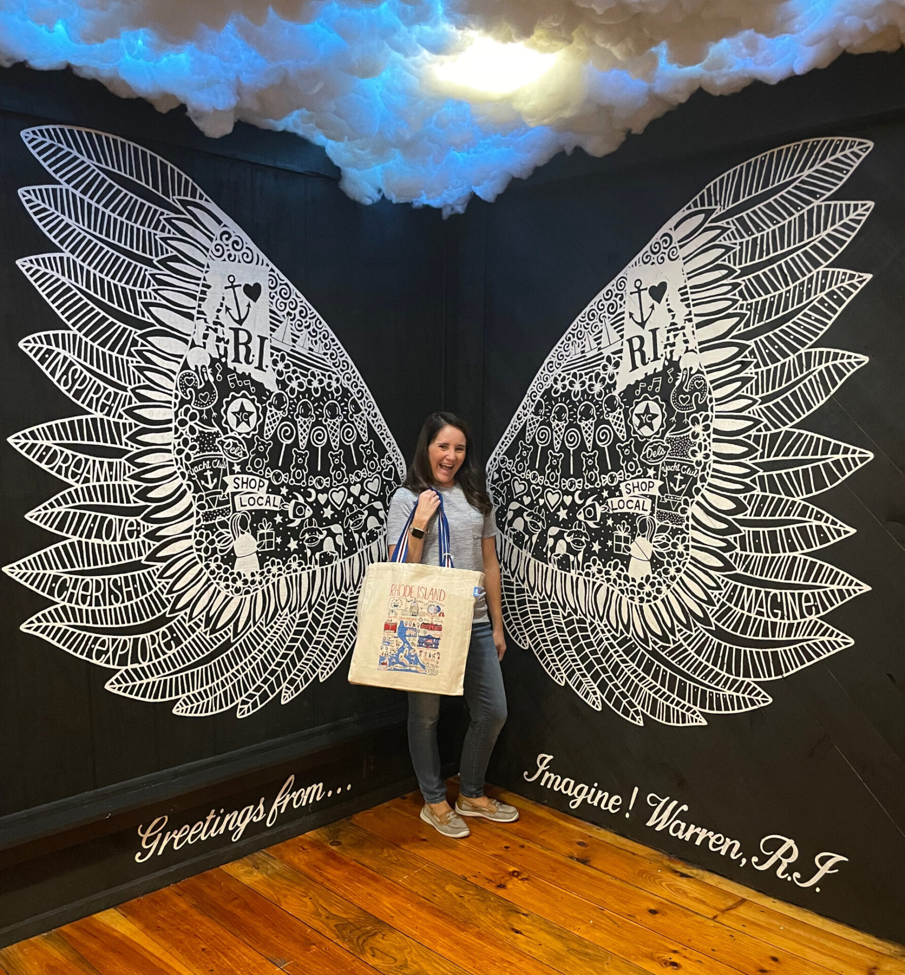 A woman poses in front of a mural with two white wings on either side protruding on the background. She is holding a bag. There are model clouds overhead.