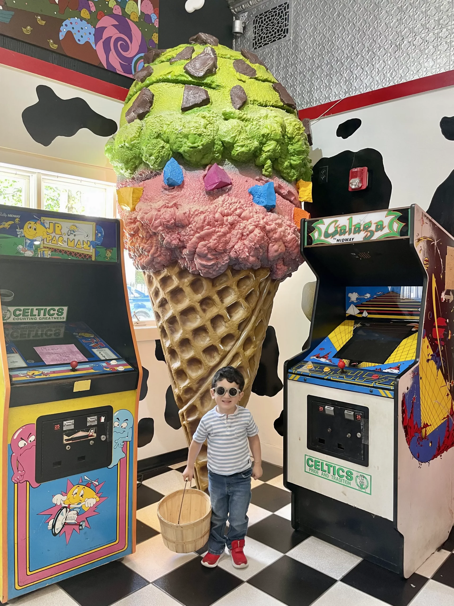 A boy stands in front of a giant ice cream cone model with two arcade machines on either side of him.